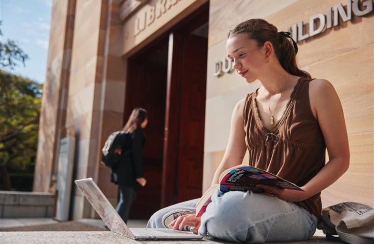A young woman sits outside the UQ Library, looking at her laptop