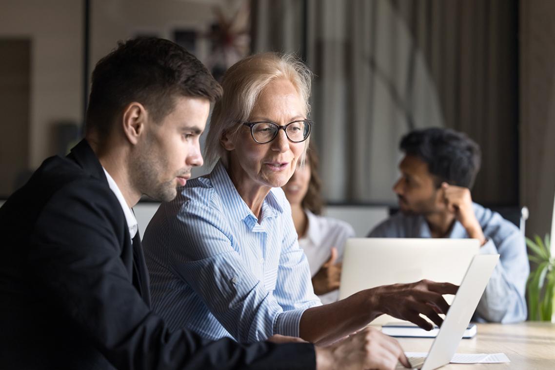 A group of people sit together at a table in a modern meeting room. In the foreground, two people sit side‑by‑side, both looking at a laptop while one of them points at the screen. Papers and a pen are on the table in front of them. In the background, two additional people sit at a separate table, each working on a laptop.