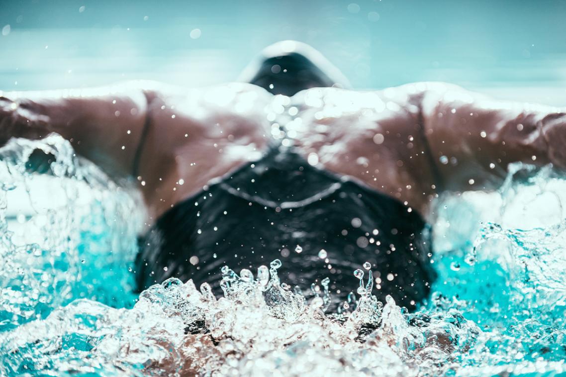 A swimmer in action doing the Butterfly stroke. Shot from behind, focus on water ripple behind the swimmer.