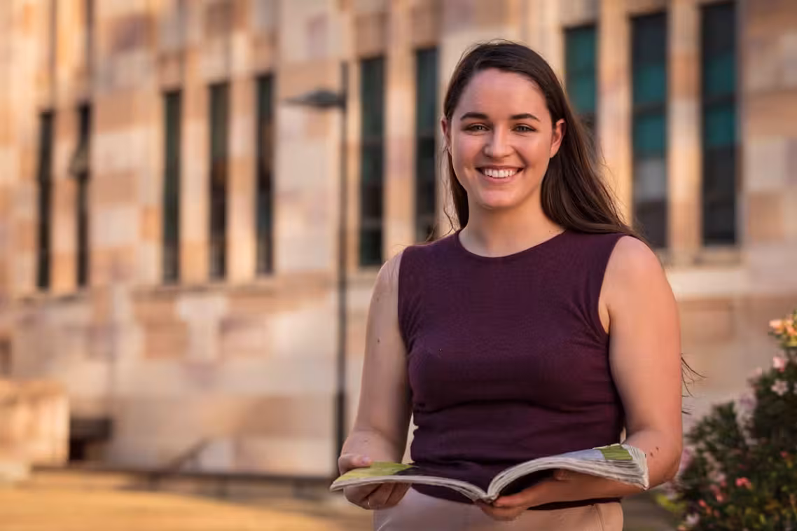Woman standing in front of a sandstone building holds an open book