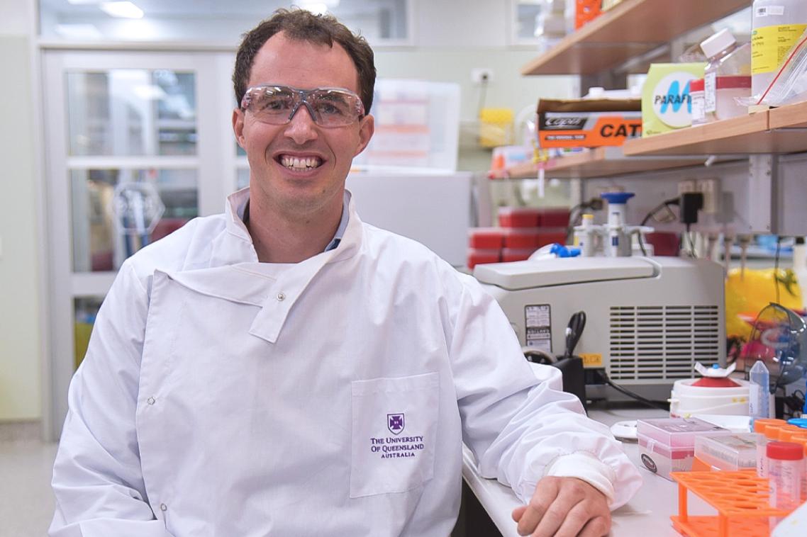 A male researcher wearing a white lab coat and safety glasses sitting in a research lab.