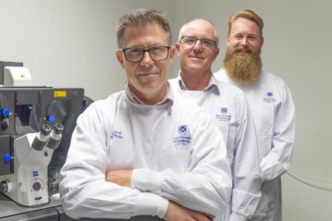 Three men in white lab coats stand next to a microscope. 